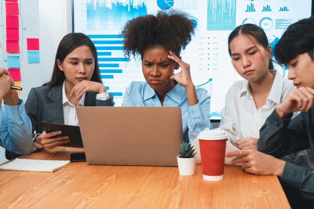 Diverse group of professionals discussing strategies for Google Business Profile management and local SEO, focused on laptop screen with data visuals, expressions of concern and concentration, office setting with coffee cup and notepad.