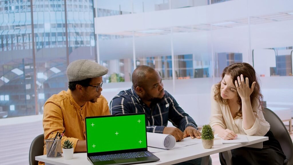 Three professionals collaborating at a modern office table, discussing strategies for Google Business Profile management, with a laptop displaying a green screen and a small potted plant.
